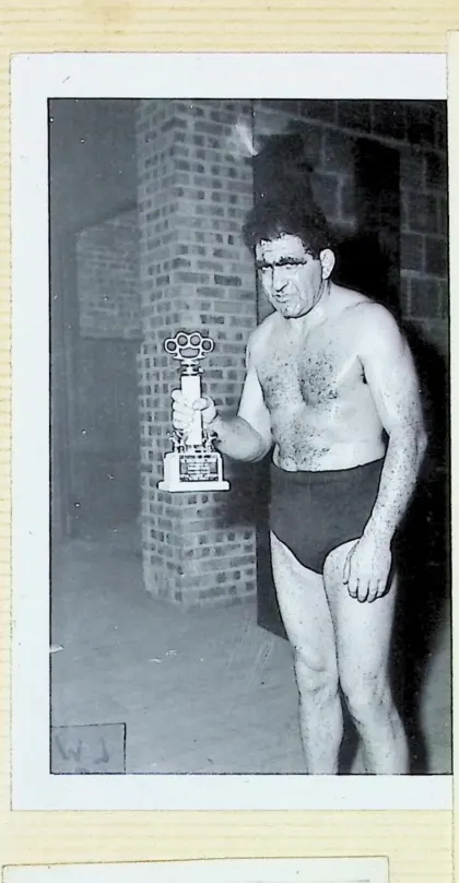 Bull Curry shirtless in wrestling trunks holding a trophy topped with a set of brass knuckles