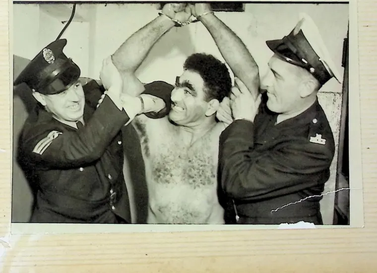 Family album photo, Bull Curry shirtless with his cuffed hands raised, flanked by two grinning police officers, Curry grinning widest of all