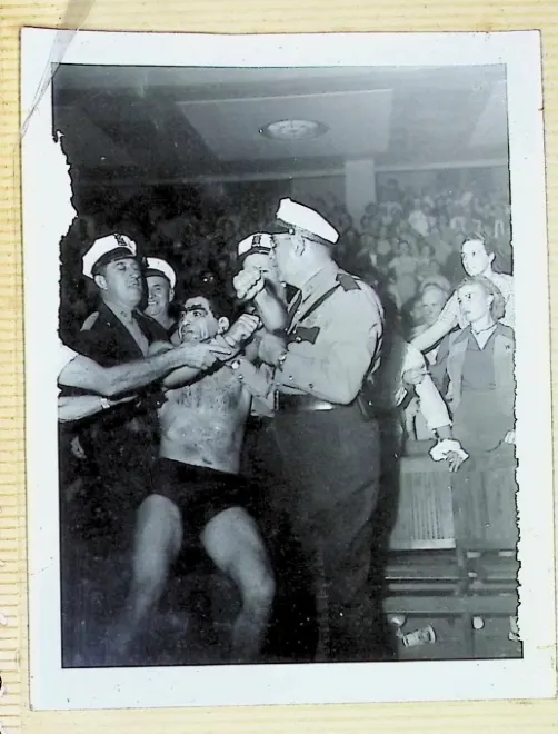 Family album photo, uniformed officers restraining a shirtless Bull Curry at ringside with an arena crowd behind them